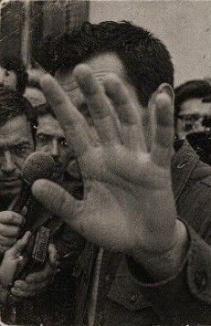 Fotografía antigua de un hombre que tapa el objetivo con la mano, rodeado de periodistas con micrófonos